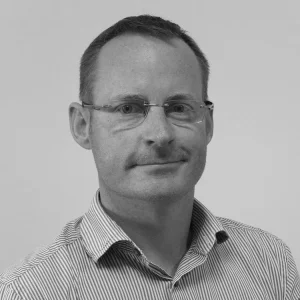 Black and white portrait of a man in glasses and a striped shirt, smiling at the camera. 