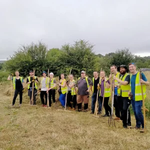 Team of volunteers in bright safety vests holding rakes and pitchforks while standing in a row on a mown, grassy area with bushes behind them.