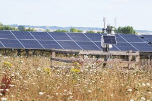 A small fenced-off weather or monitoring station with solar panels stands in a wildflower meadow, with rows of solar panels and a distant countryside landscape behind.