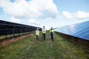 Three people in high-visibility vests walking along a grassy track between two long rows of solar panels, under a blue sky with scattered white clouds.