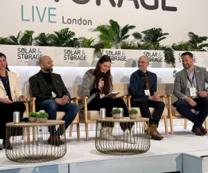 Five conference speakers smiling and seated on a stage with 'Solar & Storage LIVE London' branding and potted plants behind them. 