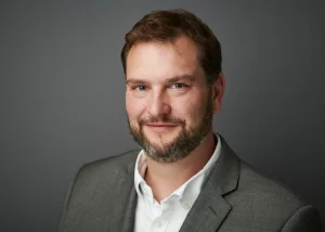 Mark is in a grey suit and white shirt posed against a plain grey background, smiling at the camera.