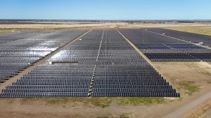 Aerial view of an extensive solar farm with countless solar panels arranged in large rectangular grids, situated on flat, dry land under a clear blue sky.