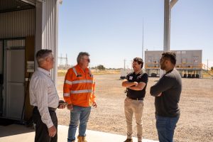 Four men stand conversing at the entrance to a building on an industrial site; one wears an orange high-visibility jacket, while the others wear business casual or dark clothing. Industrial equipment and clear blue sky are visible in the background.