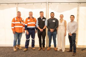 Six people stand in a row posing for a group photo inside a tent with a white backdrop. Two are wearing orange high-visibility work jackets, one is dressed in business casual with a sleeveless white vest and trousers, while the others are in dark sweaters or shirts and trousers.