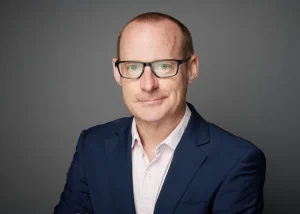 Tim is in a dark blue suit and white shirt against a plain grey background, with his face blurred for privacy.
