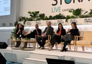 Five panelists seated on stage with microphones, plants, and 'Solar & Storage LIVE London' branding in the background, participating in a conference discussion.